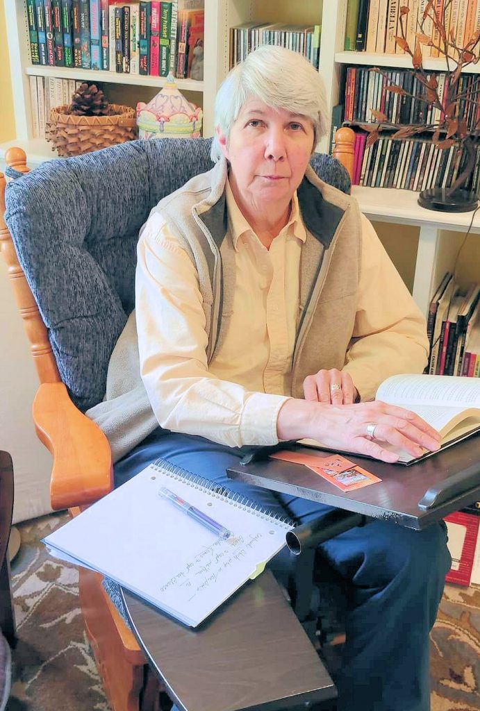 Sharon Lee sitting in blue rocker reading book on stand, notebook and fountain pen at her right hand. Photo by Steve Miller